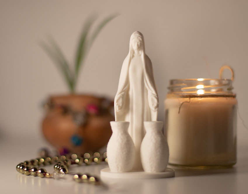 A white porcelain statue of Mary with two jars from the Wedding Feast at Cana, next to a rosary and a jar candle